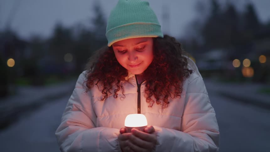 A teenage girl stands in the middle of the road wearing a winter jacket and hat looking down at a light shining on her arm with a smile on her face at sundown