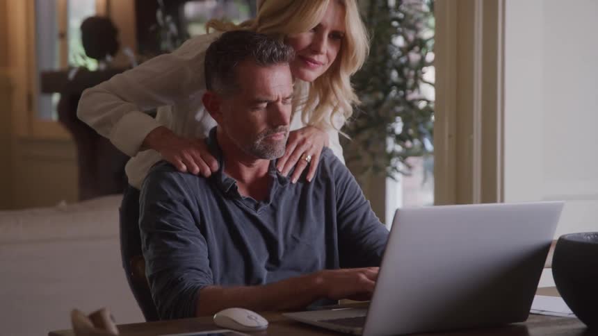 The video shows a man working on a laptop at a table a woman walks up behind him and gently places her hands on his shoulders then rests her head on his shoulder