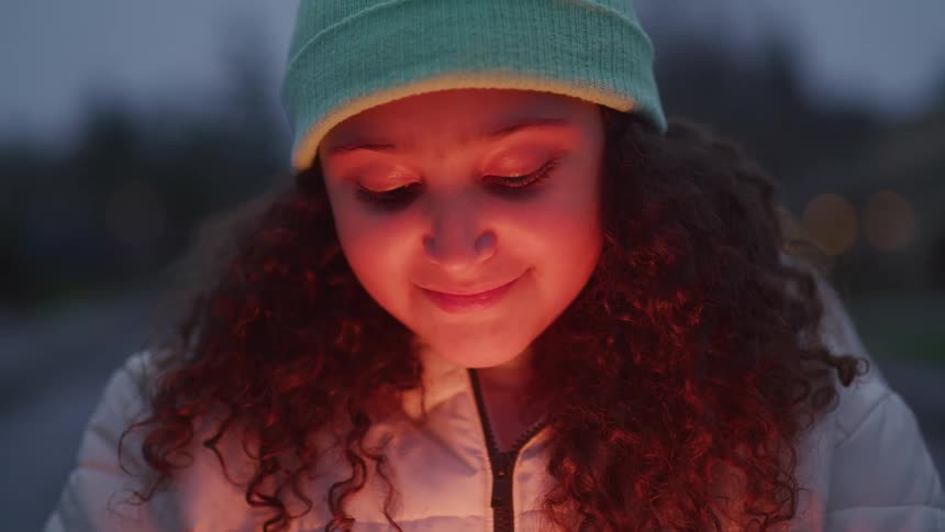 The video shows a young person with curly hair and a beanie looking down at something held in their hands the light from the object illuminates their face and hair