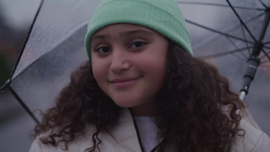 The video shows a young girl with curly hair and a beanie holding a clear umbrella in the rain she looks at the camera and smiles appearing happy despite the weather