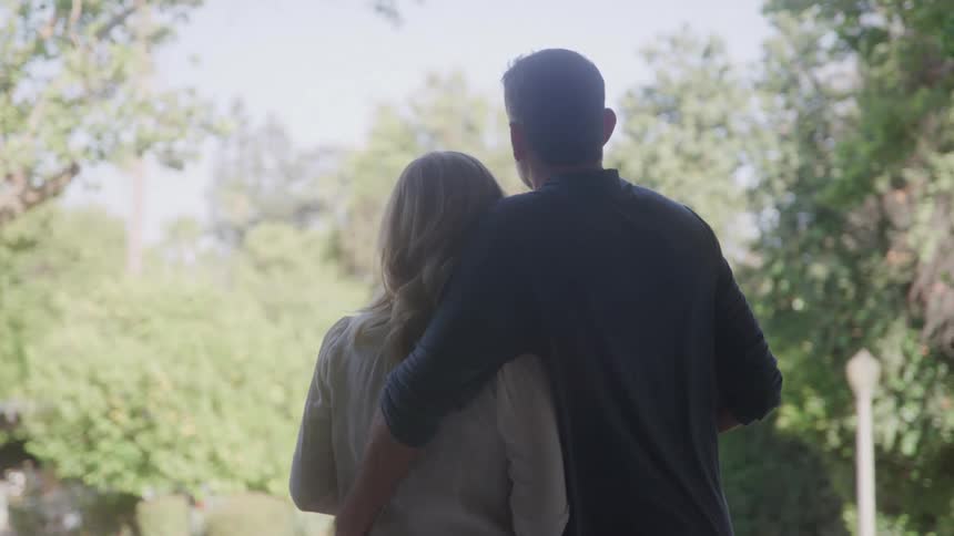 The video shows a back view of a man and woman standing together in front of a window the man has his arm around the womans shoulder as they look out at the greenery outside