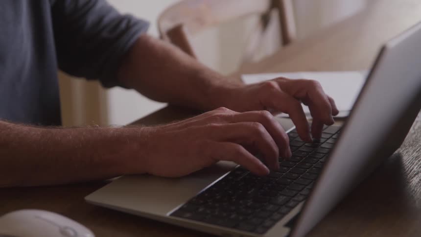 The video shows a man with a beard wearing a dark gray polo shirt sitting at a table and working on a laptop computer he appears focused looking down at his screen and typing on the keyboard