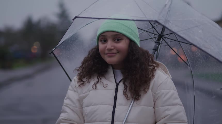A young girl with a gentle smile and curly hair stands on a wet street holding a clear umbrella above her head