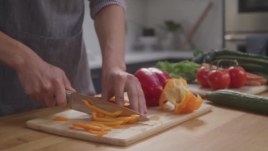 A person slices yellow bell peppers beside fresh vegetables on a wooden board