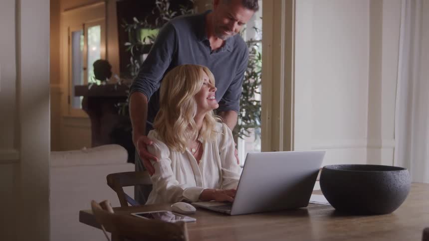 A woman types on a laptop at the dining table as a man stands nearby watching her rubbing her shoulders