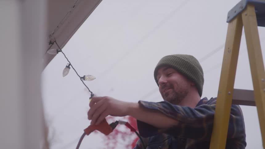 A man on a ladder hangs string lights along the roof while smiling