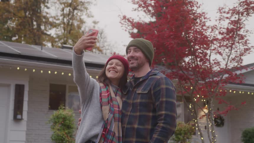 The video shows a man and woman standing outside a house taking a selfie together they are both smiling and wearing hats and scarves suggesting it is a cool day