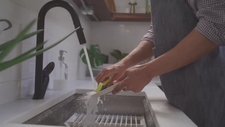 The video shows a persons hands washing a white plate with a yellow sponge the water is running from a black faucet into a sink
