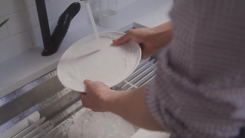 The video shows a persons hands washing a white plate with a yellow sponge the water is running from a black faucet into a sink