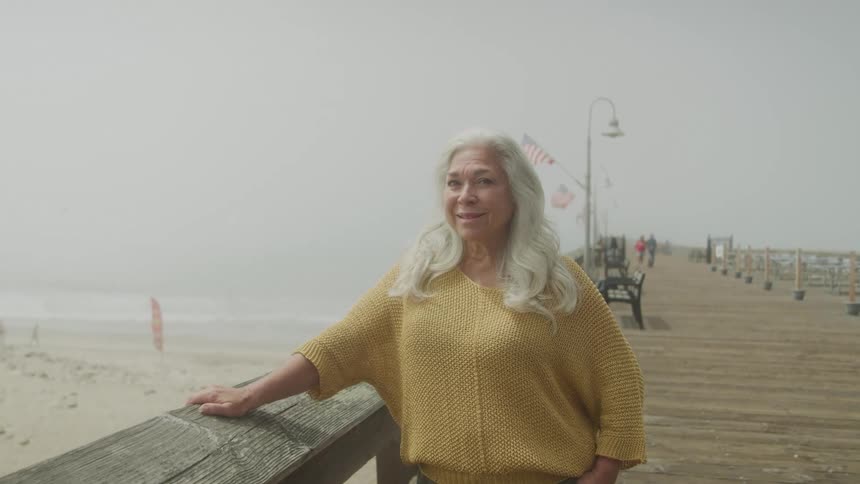 An older woman with long gray hair stands on a foggy pier leaning on a wooden railing and smiling warmly at the camera