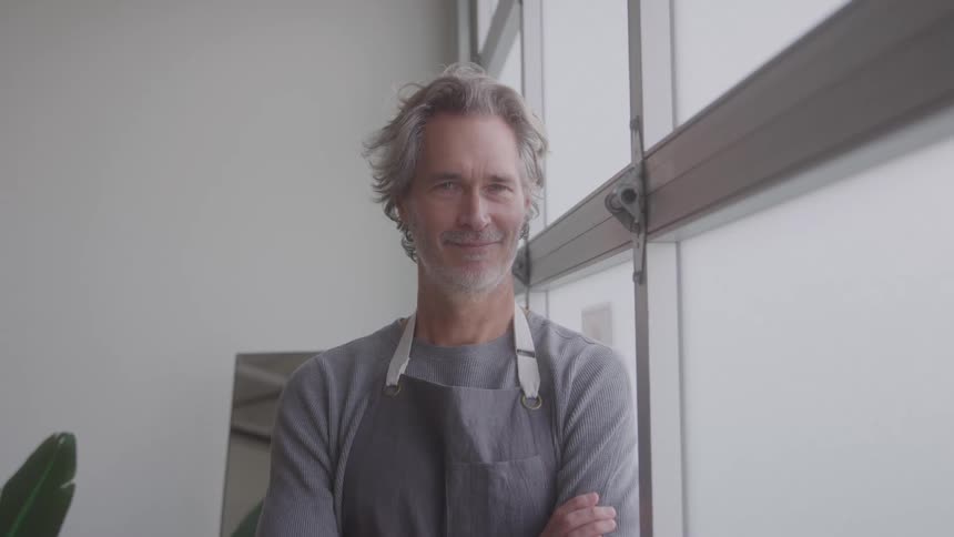 The video shows a middleaged man with graying hair and a beard wearing a gray apron over a dark gray shirt he stands with his arms crossed looking directly at the camera with a friendly smile