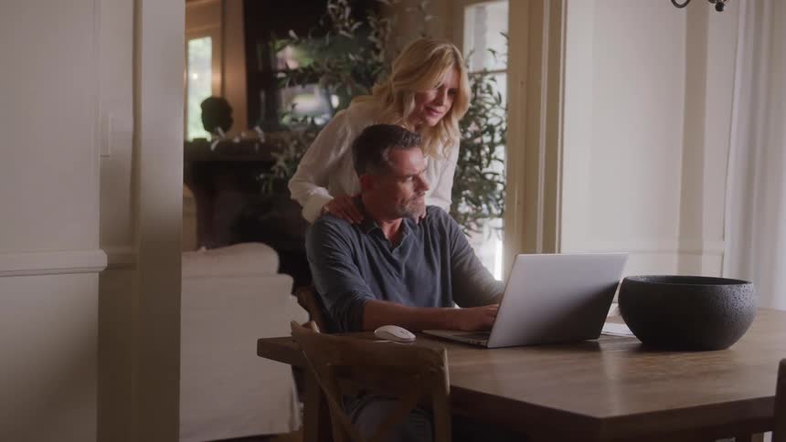 A woman walks into the dining room where her husband is seated working on a laptop at midday