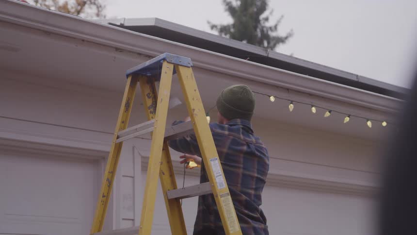 A man wearing a beanie and a plaid shirt stands on a yellow ladder focused on the task of hanging string lights on the gutter of a house