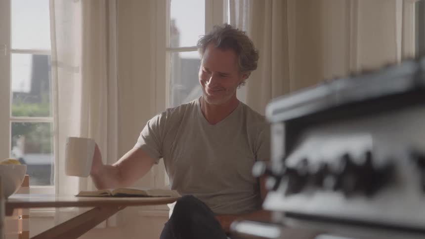A man sitting at a coffee table drops a coffee mug onto it and continues reading the open book in front of him indoors at midday