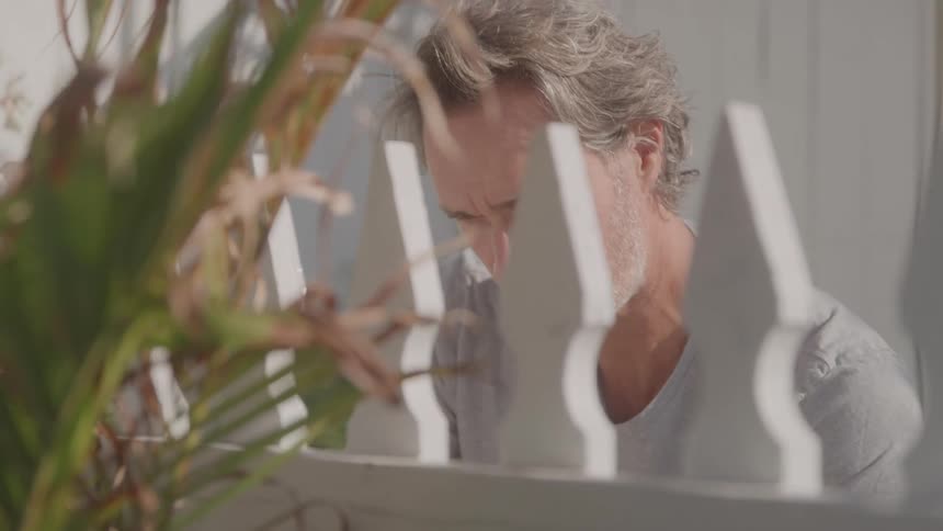 The video shows a man with graying hair and a beard wearing a gray tshirt who is painting a white picket fence he is focused on his work painting carefully with a brush