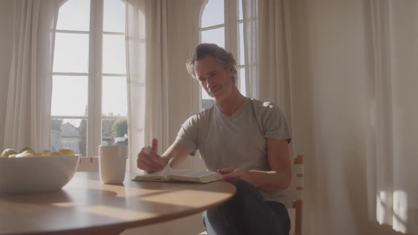 A middleaged man sits at a sunlit table smiling and turning the pages of a book while enjoying a peaceful moment