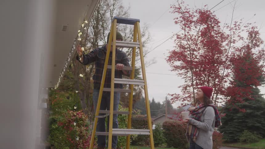 A man on a ladder sets up christmas lights on the roof of his home while his wife holding a coffee cup stands beside him watching at midday with trees surrounding the house