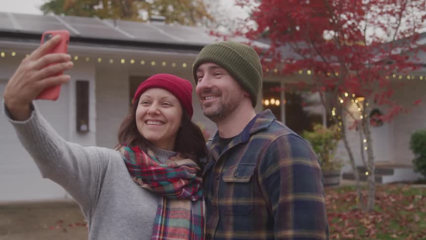 A joyful couple in winter hats and scarves smiles as they take a selfie together in front of a house decorated with holiday lights