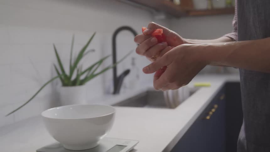 A chef in the kitchen picks up some sliced tomatoes from the chopping board and places them into a bowl on a scale