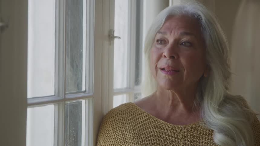 The video shows a closeup of an elderly womans face with long gray hair she is looking out a window with a serious or thoughtful expression