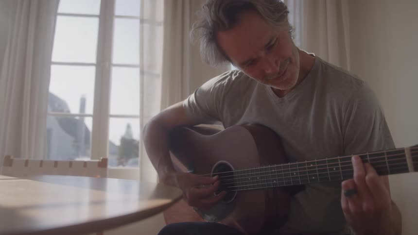 A middleaged man sits in his dining room playing a guitar and smiling at midday