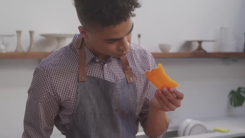 A young boy standing in the kitchen picks up a piece of produce and holds it near his nose to smell it at midday