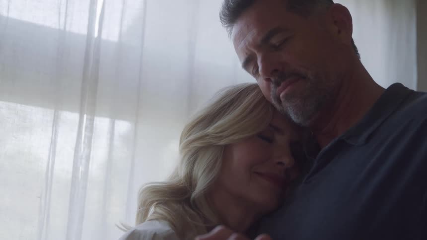 A loving middleaged couple slow dances in a sunlit room embracing each other tenderly with their eyes closed