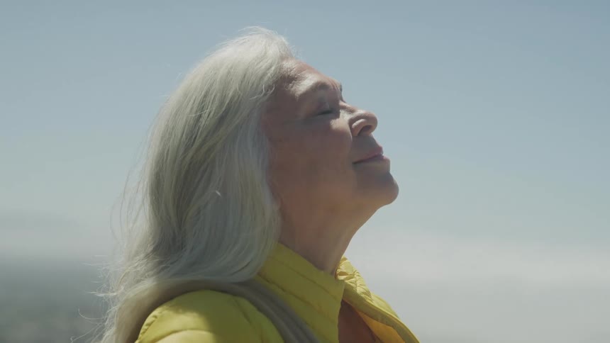 The video shows a closeup of an elderly woman with long gray hair she is wearing a yellow shirt and looking up with her eyes closed as if enjoying the sun on her face