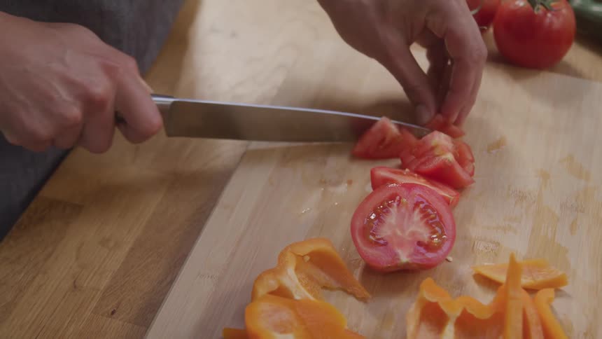Hands slice tomato on a wooden cutting board next to orange bell pepper strips