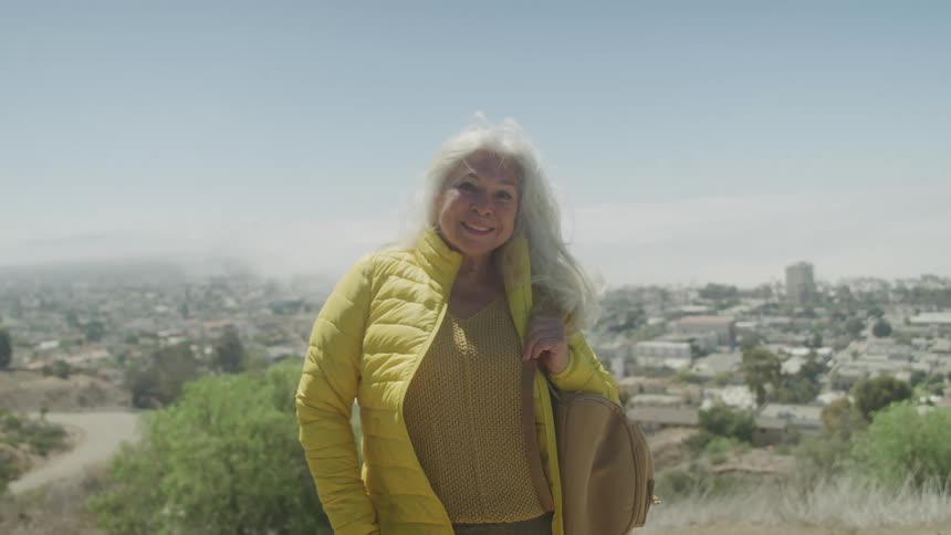 An elderly woman wearing a jacket and holding a handbag stands on a hill surrounded by grass on the outskirts of the city at midday