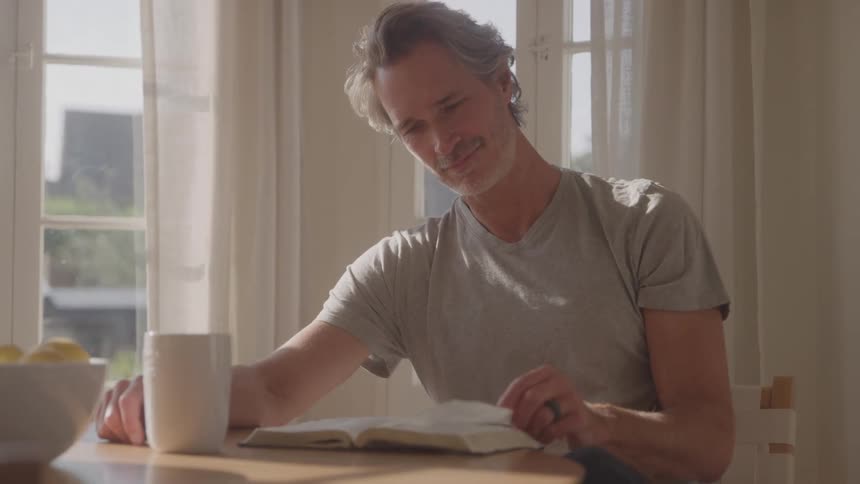 A middle aged man flips and reads through a book while sitting at a dining table indoors during daytime