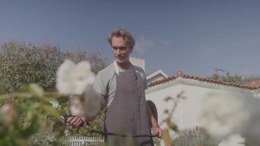 The video shows a man with a beard and graying hair wearing a gray shirt and apron standing in a garden he is holding a hose and looking down as if he is about to water the plants
