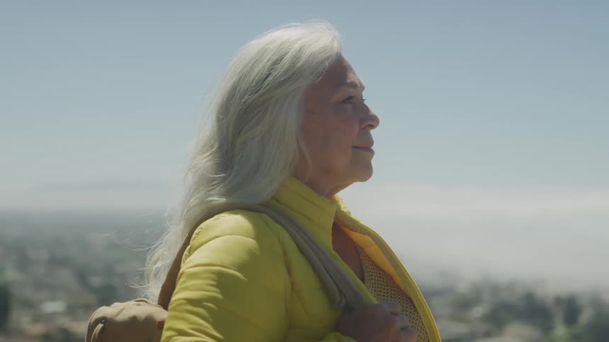 A side profile shot shows an older woman with long gray hair standing on a hilltop looking out at a cityscape below she is wearing a yellow jacket and carrying a bag over her shoulder