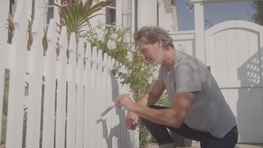 A middleaged man paints his fence with a brush and white paint at midday smiling