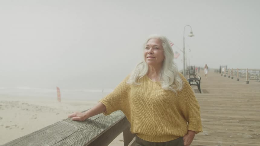 An elderly woman with long gray hair stands on a bridge wearing a yellow jacket and smiling