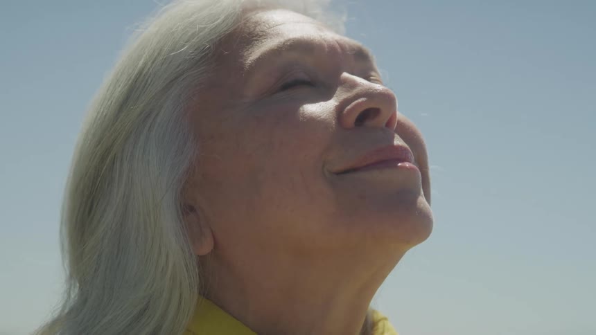 A woman with long gray hair tilts her face toward the sun with closed eyes and a peaceful smile