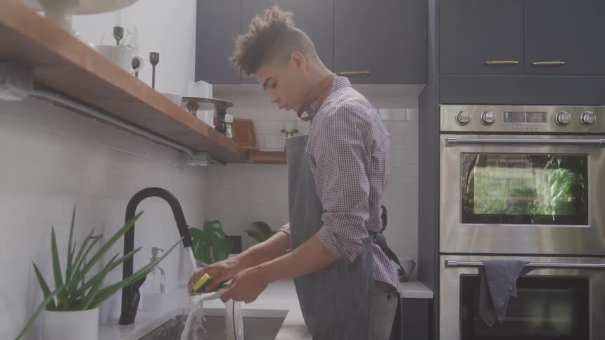 A teenage boy cleans and washes a plate in the kitchen sink at midday