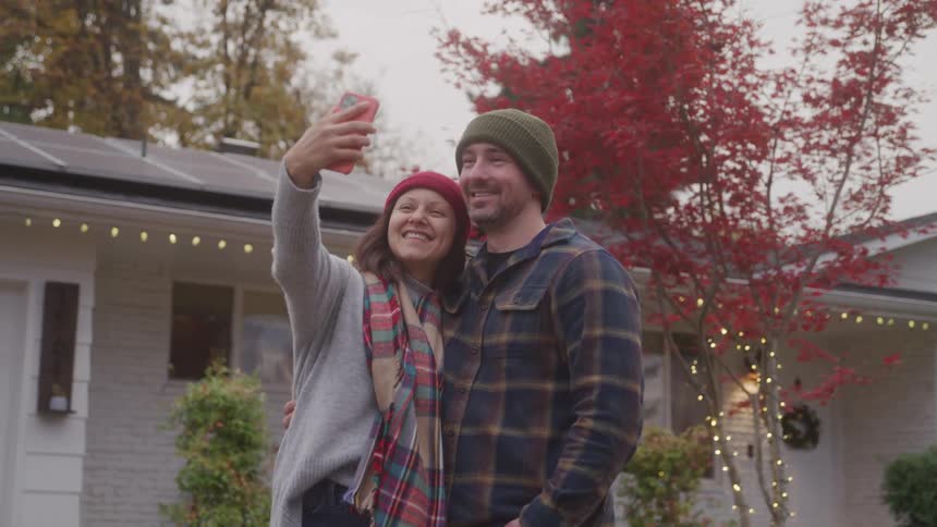 Couple in winter clothes takes a smiling selfie in front of a decorated house