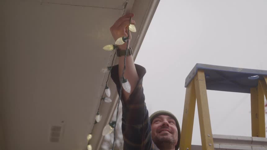 The man smiles while hanging christmas lights from the roof