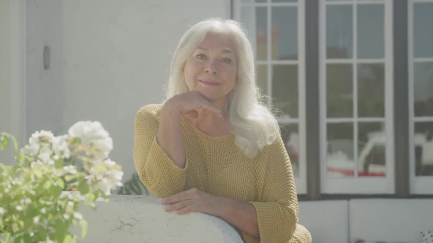 An elderly woman smiles while leaning on a porch railing