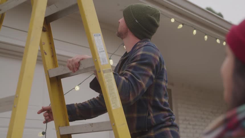A young man installs festive lights around a building roof with a ladder outdoors during daytime