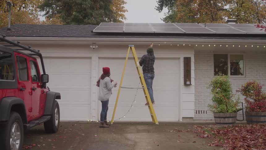 A woman on the ground and a man on a yellow ladder work together to hang white christmas lights on the front of a house while a red jeep is parked nearby