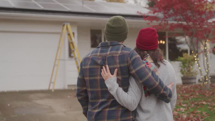 A couple hugs each other while standing in their driveway staring at their house with a ladder in front of the closed garage door and a series of solar panels on the roof at midday