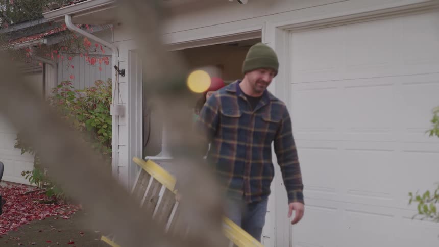 A man and a woman walk out of a garage and the man carries a yellow ladder while the woman carries a large container of christmas decorations
