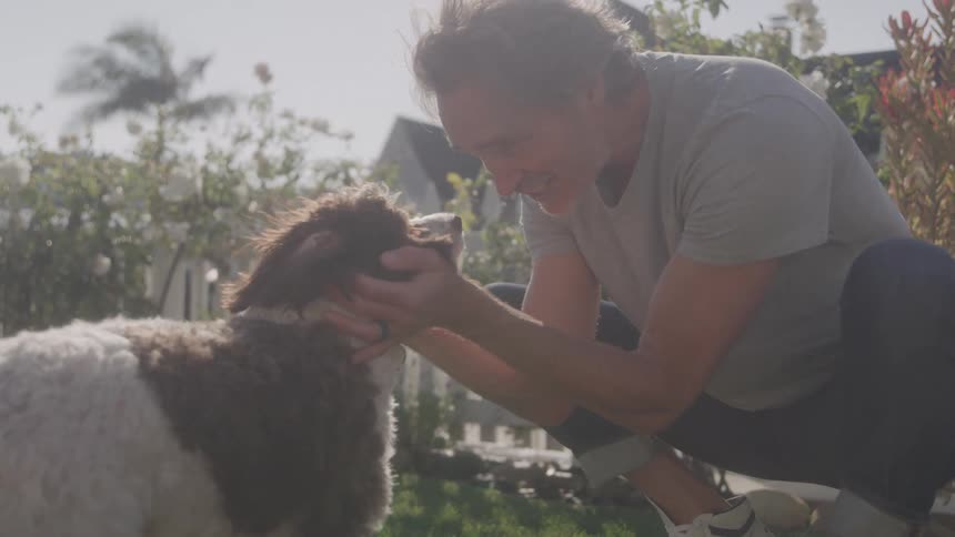 Man crouches smiling while petting a dog in the garden