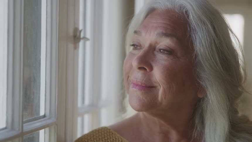 A woman with long gray hair gazes out a window with a peaceful smile in soft natural light