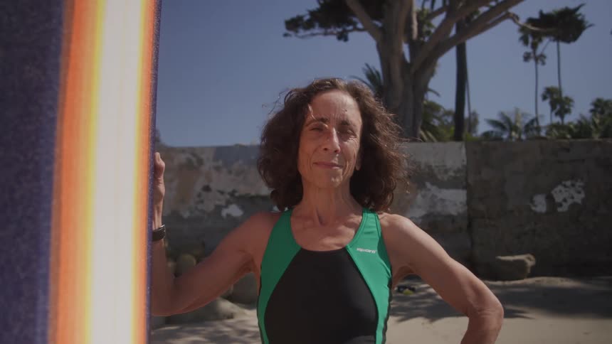 A woman with long brown curly hair and a black and green one piece swimsuit stands outdoors next to a surfboard looking directly at the camera