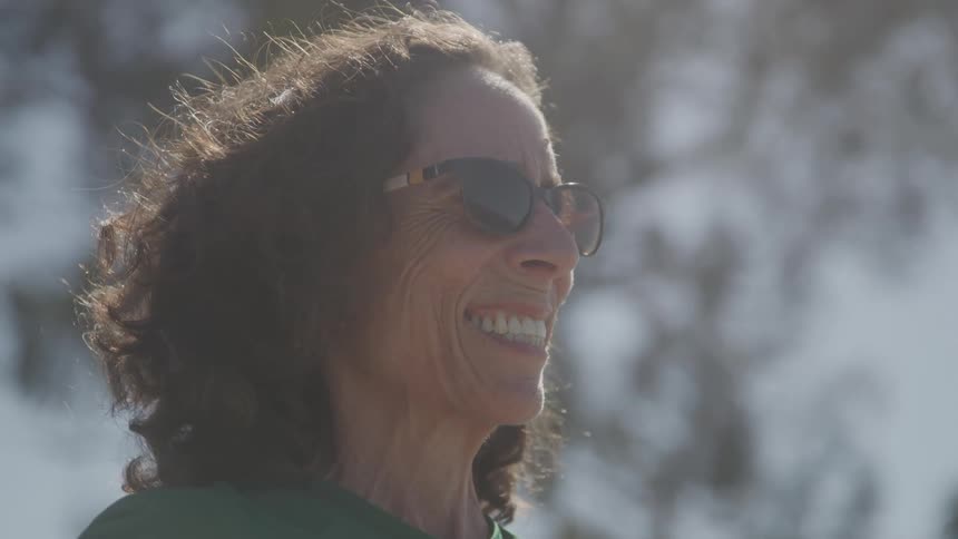 The video shows a closeup of a smiling woman with dark curly hair and sunglasses the sun is shining brightly from behind her giving her a glowing appearance