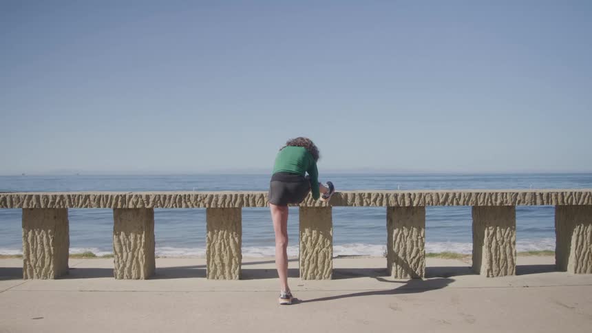 A woman in a green top and black skirt is stretching with her leg propped up on a low concrete wall overlooking the ocean on a sunny day