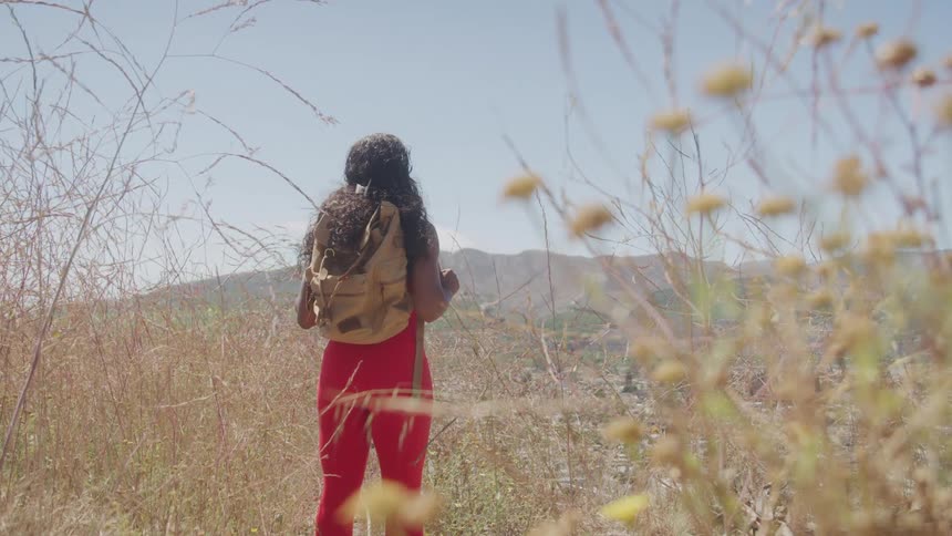A young black woman carrying a backpack while standing outdoors in a grassland area during daytime